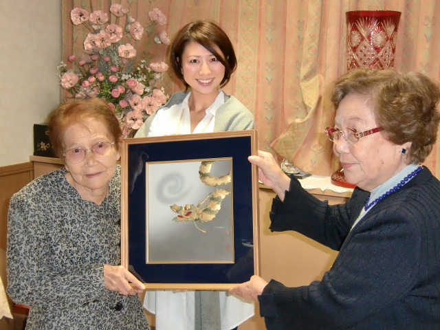 田倉理事長（右）に「辰｝の押絵を手渡す稲葉さん（左）（向こうは孫娘の友希さん）